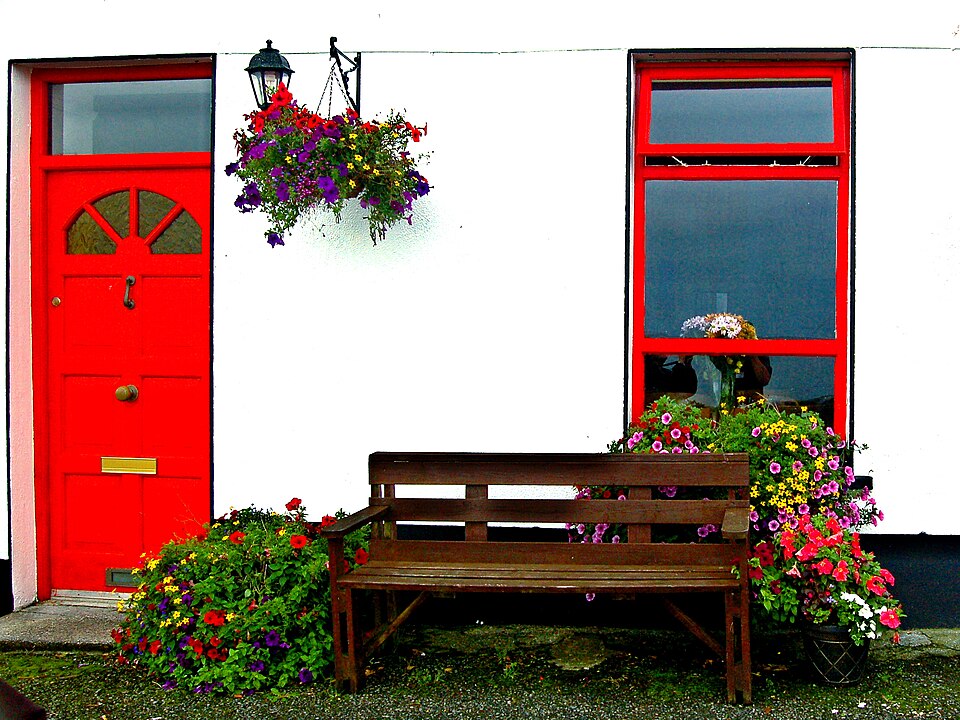 Red door beside a bench and flower planters at a whitewashed bed-and-breakfast.